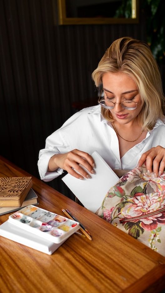 Camila Teixeira desenhando uma nova peça no caderno, com lápis de cor, retalhos de tecido e uma xícara de café ao lado, no ateliê de Ipanema.